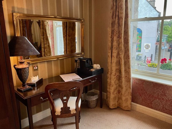 A hotel room desk with a mirror, lamp, and coffee maker by a window.