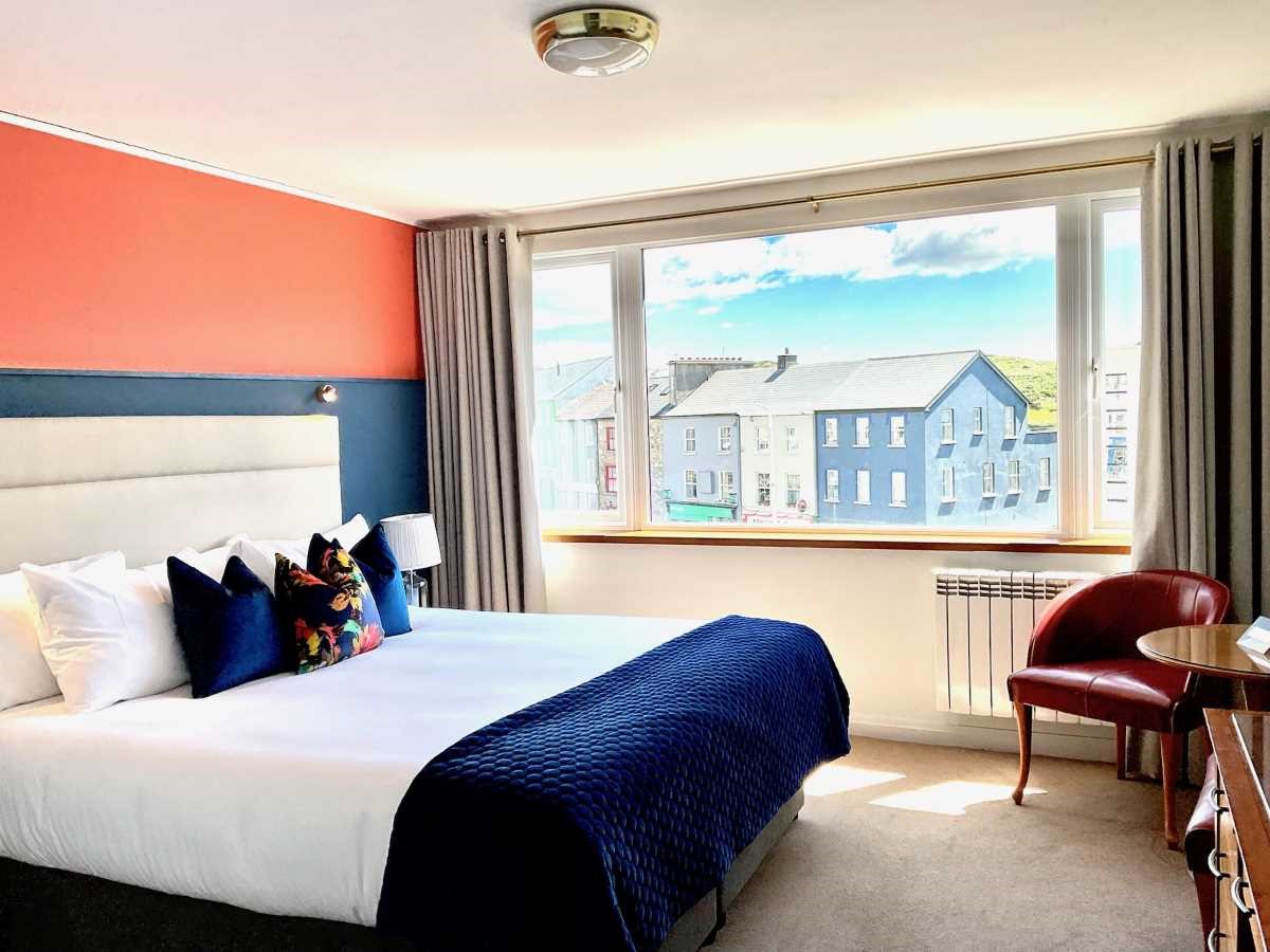 Bright hotel room with a large bed, colorful accents, and a window view of houses.